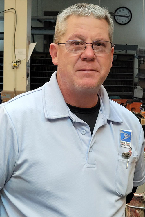 Smiling man in postal uniform stands on workroom floor
