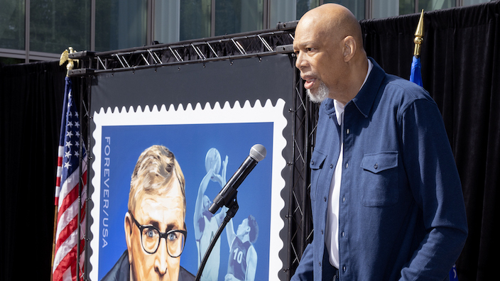 Kareem Abdul-Jabbar speaks at a podium near a poster displaying a stamp that shows John Wooden and two players reaching for a basketball in midair