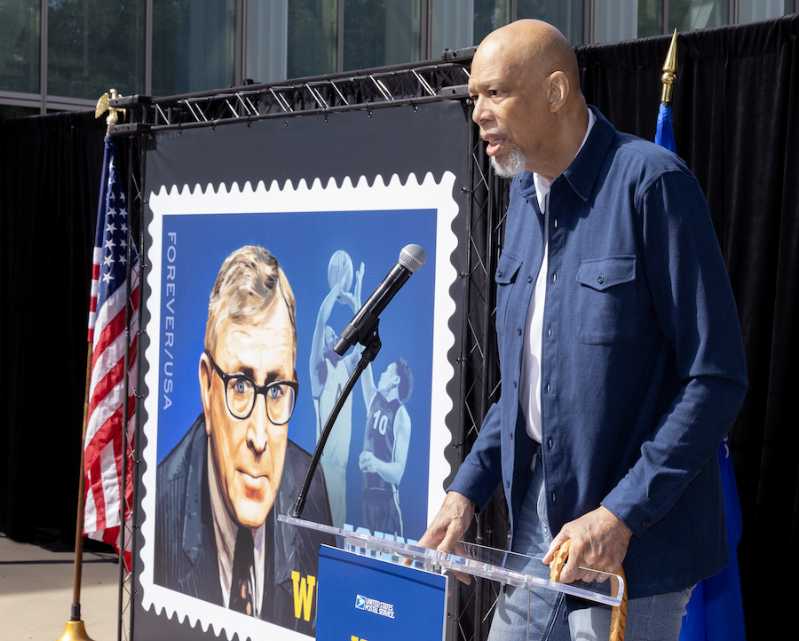 Kareem Abdul-Jabbar speaks at a podium near a poster displaying a stamp that shows an illustrated portrait of John Wooden