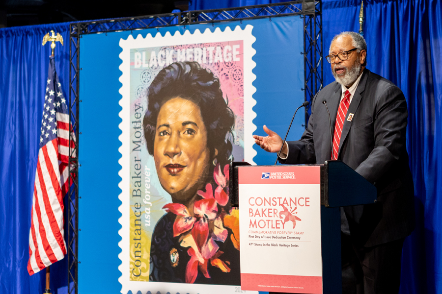 A man wearing a suit speaks at a podium near a poster showing the Constance Baker Motley stamp