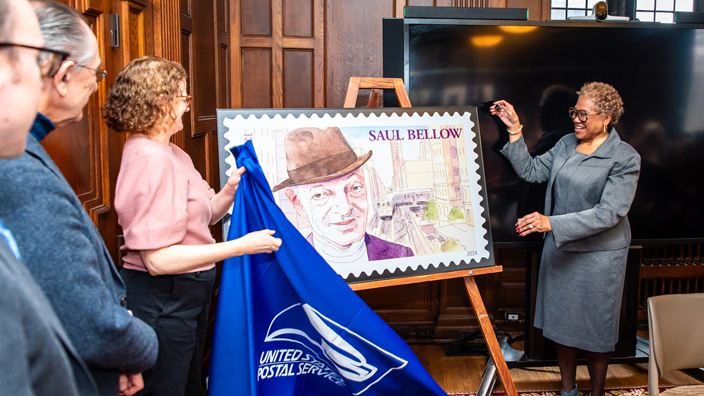 A group of women remove a cloth cover from a poster displaying an image of the Saul Bellow postage stamp