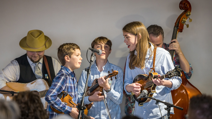 Three older children wearing light blue shirts play instruments and sing on a stage