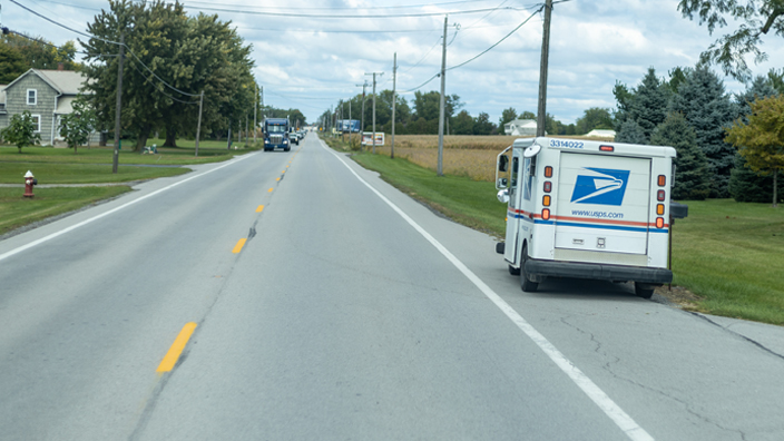 A USPS delivery vehicle is parked on the shoulder of road.