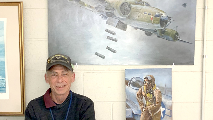 A man wearing a ball cap stands next to paintings of a World War Two bomber and an airman.