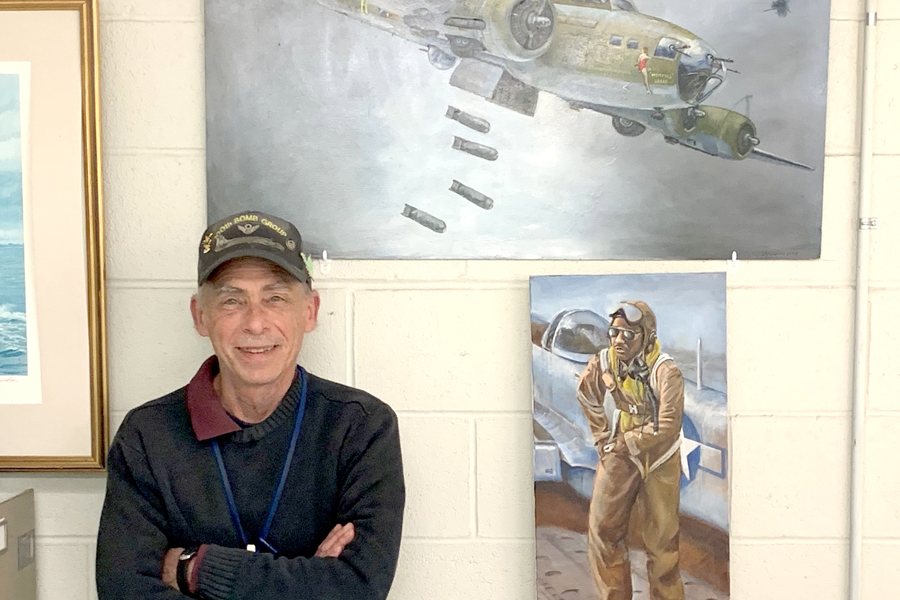 A man wearing a ball cap stands next to paintings of a World War Two bomber and an airman.