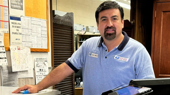 A man in a light blue USPS polo shirt stands behind a counter