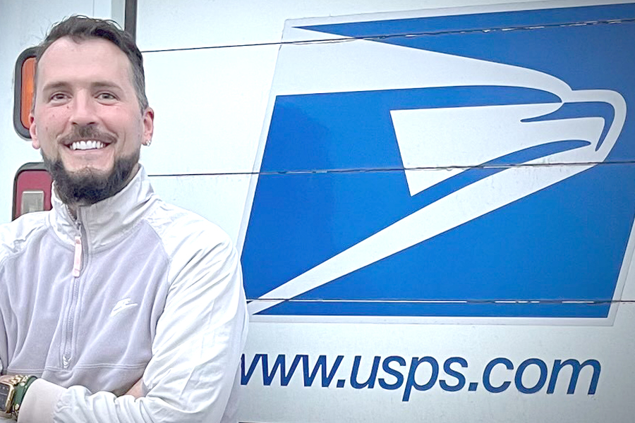 A smiling male Postal Service employee standing in front of a postal vehicle