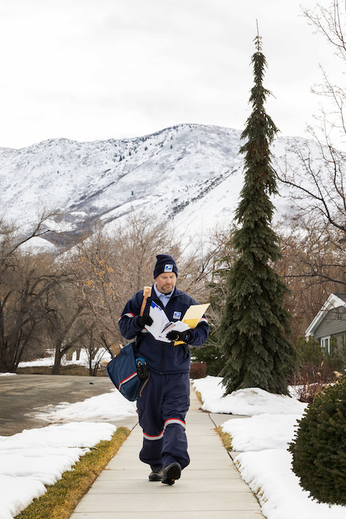 A USPS letter carriers walks along a sidewalk with a mountain in the background