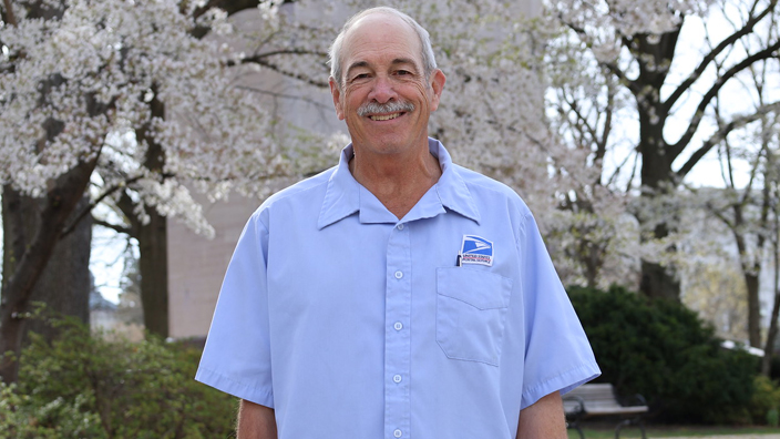 Smiling man in postal uniform stands near cherry blossom trees