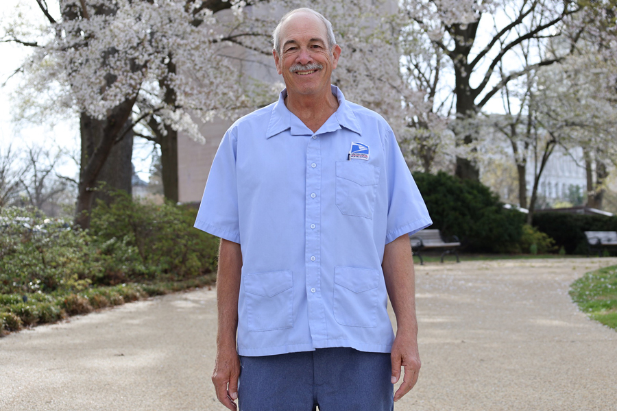 Smiling man in postal uniform stands near cherry blossom trees