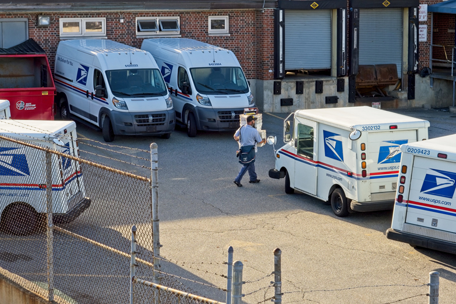 A USPS employee carries packages to a USPS delivery vehicle near a loading dock at a postal facility