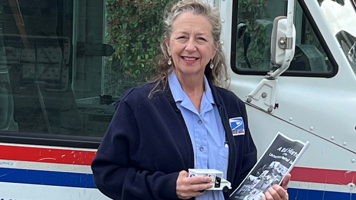 A female letter carrier holding a toy LLV and a copy of "A Kids' History of the United States Postal Service"