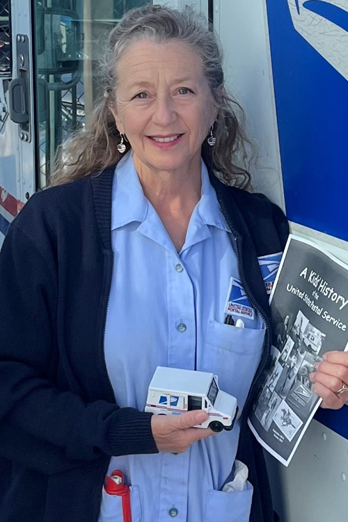 A female letter carrier holding a toy LLV and a copy of "A Kids' History of the United States Postal Service"