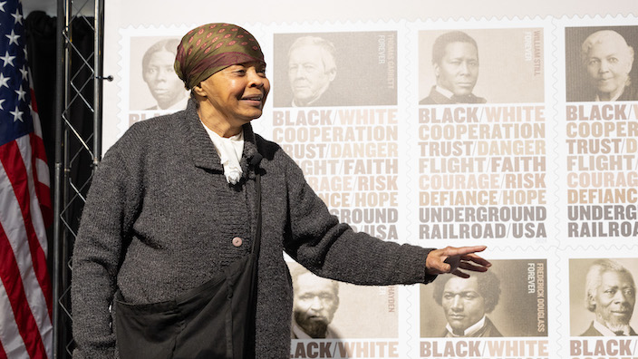 A Black woman in a Civil War era costume speaks in front of an Underground Railroad stamp poster