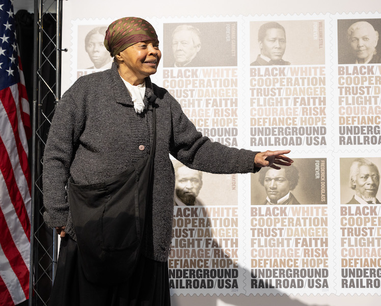 A Black woman in a Civil War era costume speaks in front of an Underground Railroad stamp poster