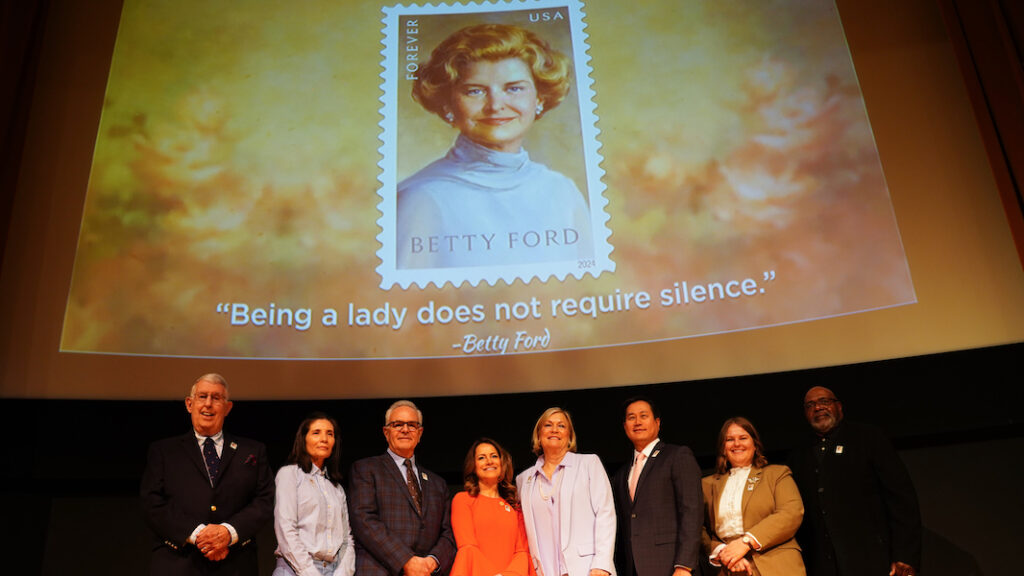 A group of smiling people stand on stage under video screen displaying Betty Ford stamp image