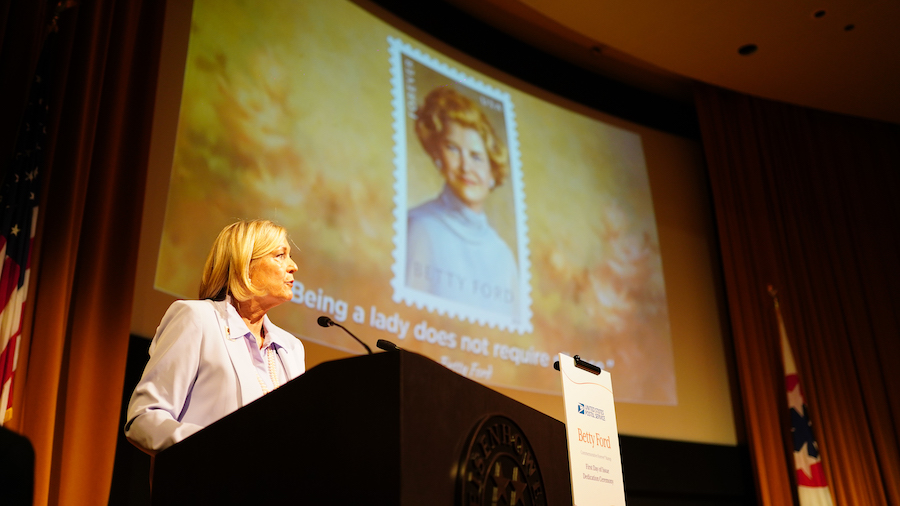Woman speaks at podium on stage near screen displaying Betty Ford stamp image
