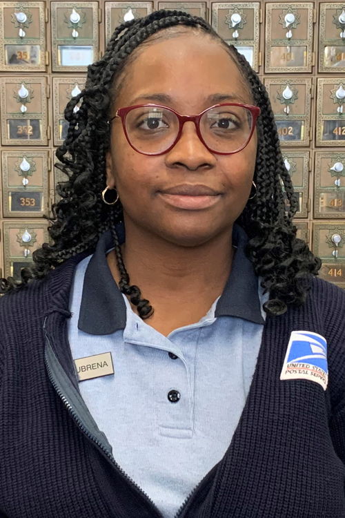 A female Postal Service employee stands in front of a wall of mailboxes