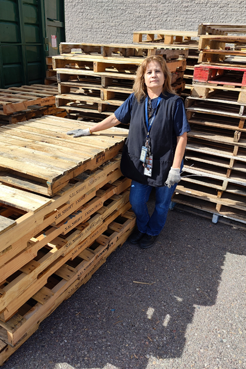 A Postal Service employee stands next to stacks of wood pallets.