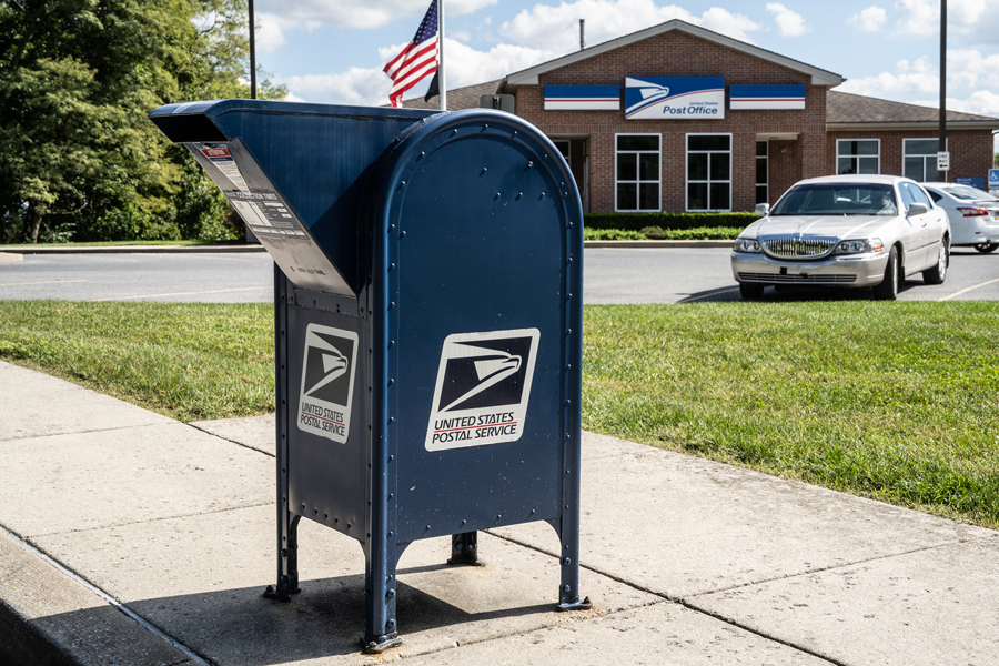 A blue U.S. Mail box in the foreground with a Post Office in the background