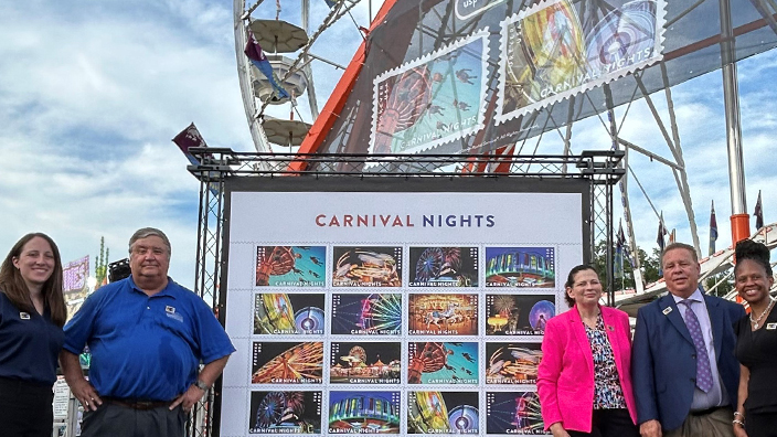 USPS officials and other guests stand on either side of a large photo of the USPS Carnival Nights stamps