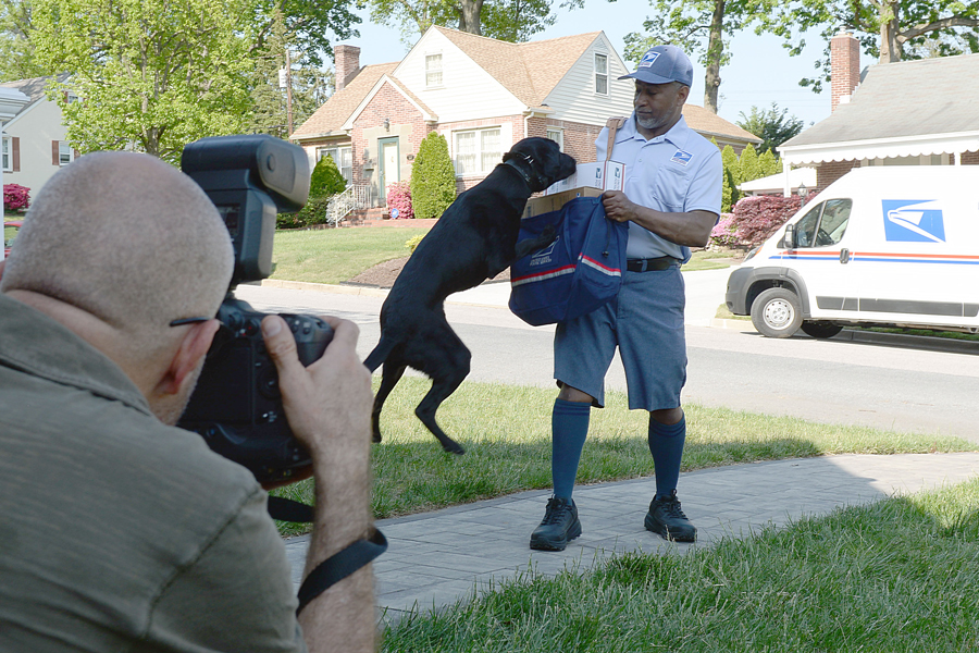 A photographer photograps a dog jumping up toward a letter carrier for USPS National Dog Bite Awareness Campaign