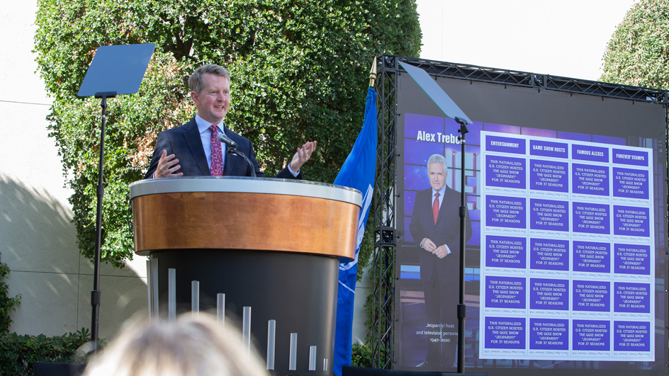 Smiling man stands at lectern near stamp poster