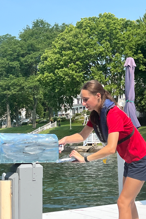 A woman places mail into a mailbox on a dock at a lake.