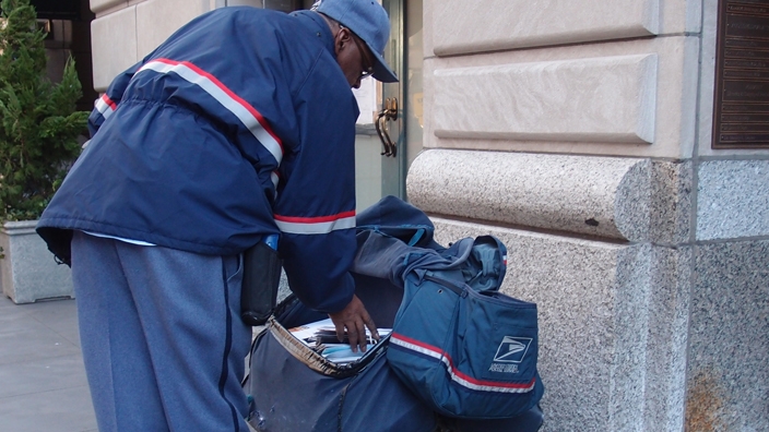 Letter carrier reaches into mail satchel while standing on sidewalk