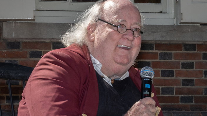 A man dressed in costume as Benjamin Franklin speaks into a microphone