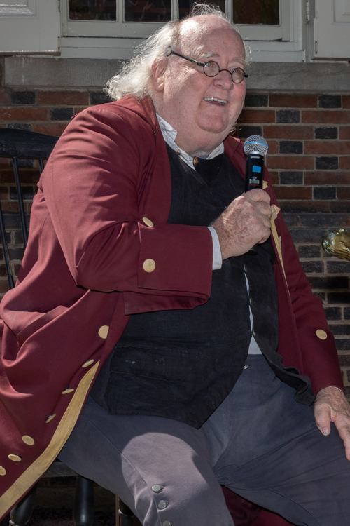 A man dressed in costume as Benjamin Franklin speaks into a microphone