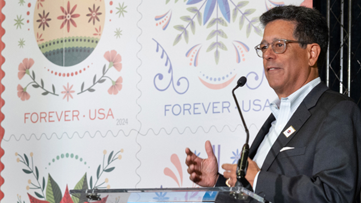 USPS art director Antonio Alcala stands at a lectern with the large-size image of the USPS Holiday Joy stamps behind him.