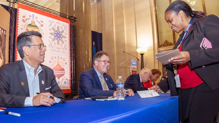 A woman stands on one side of a table while others are sitting on the opposite side of the table signing programs.