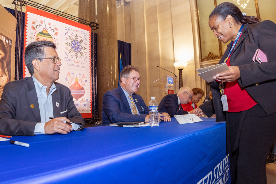 A woman stands on one side of a table while others are sitting on the opposite side of the table signing programs.