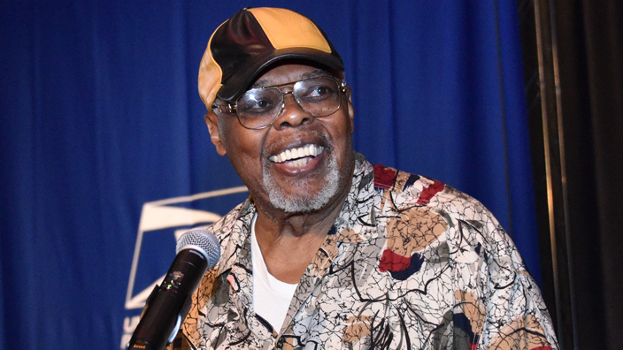 Sala Udin, wearing a cap, speaks from behind a lectern.