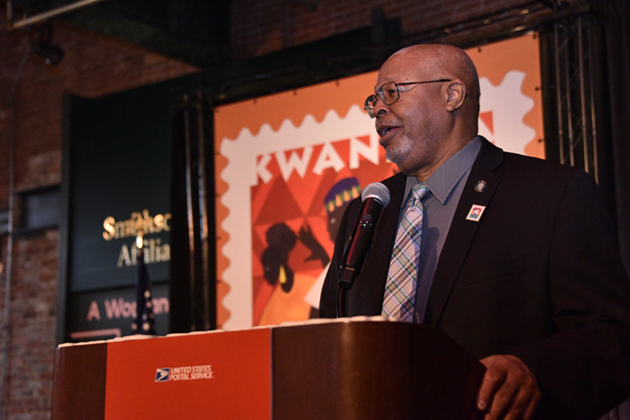 Man in suit speaks at lectern near poster displaying a Pan African-themed stamp image