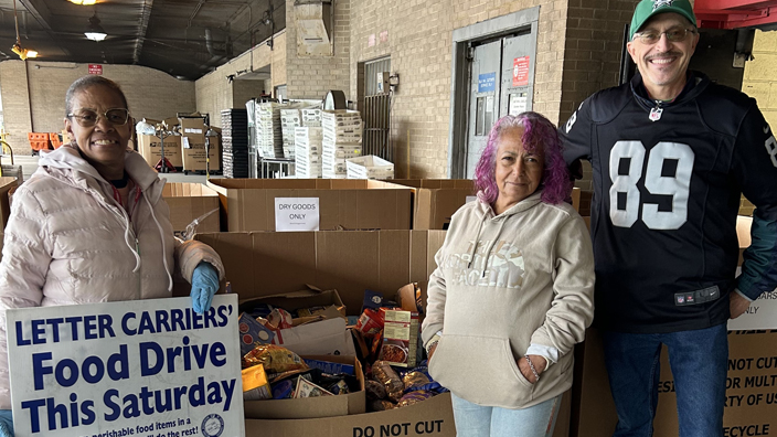 Three women stand in front of boxes of donated food items.