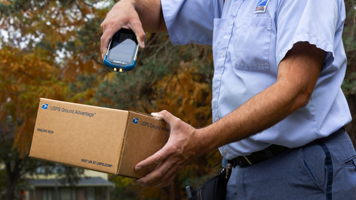 A letter carrier points a handheld scanning device at a package he delivering.