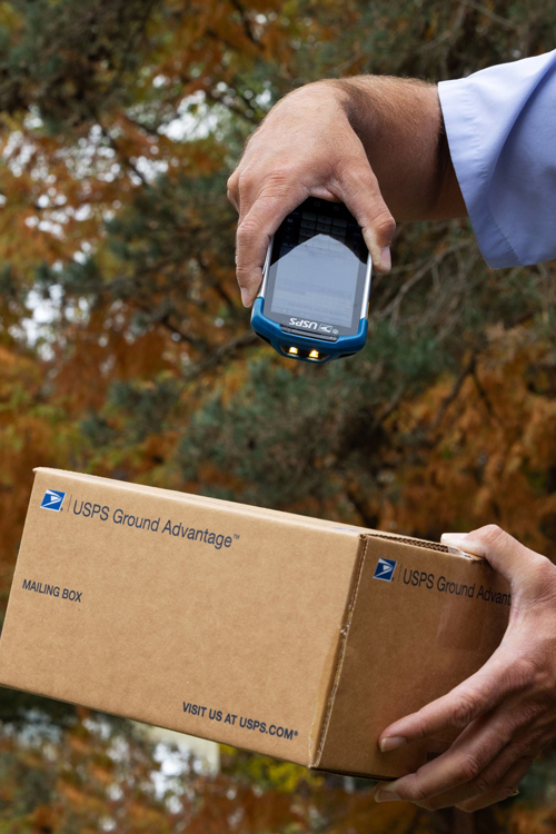 A letter carrier points a handheld scanning device at a package he delivering.