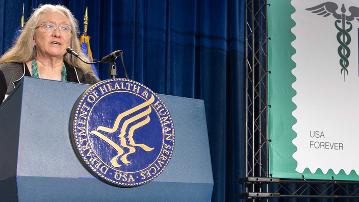 A woman with long hair speaks from behind a lectern bearing the seal of the U.S. Department of Health and Human Services near an enlarged image of the USPS Thank You, Healthcare Community stamp.