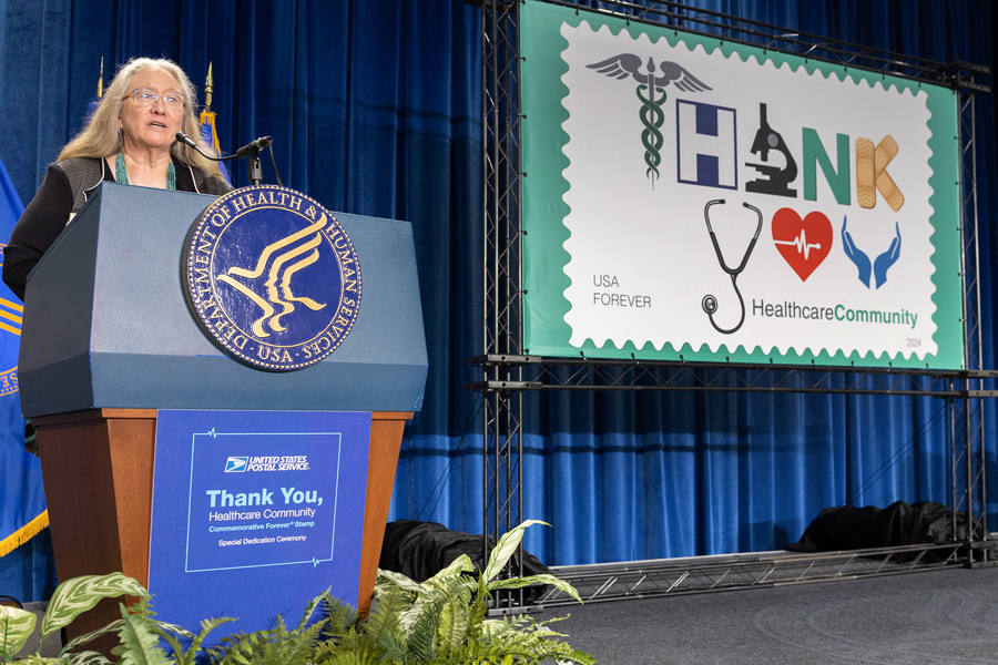 A woman with long hair speaks from behind a lectern bearing the seal of the U.S. Department of Health and Human Services near an enlarged image of the USPS Thank You, Healthcare Community stamp.