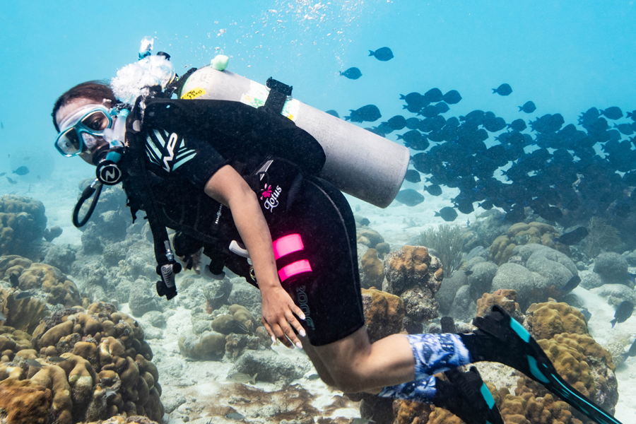 A woman swims underwater while wearing an oxygen tank, breathing mask and wetsuit.