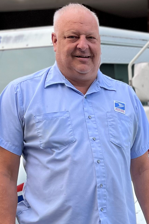 A USPS letter carrier stands in front of a USPS delivery vehicle