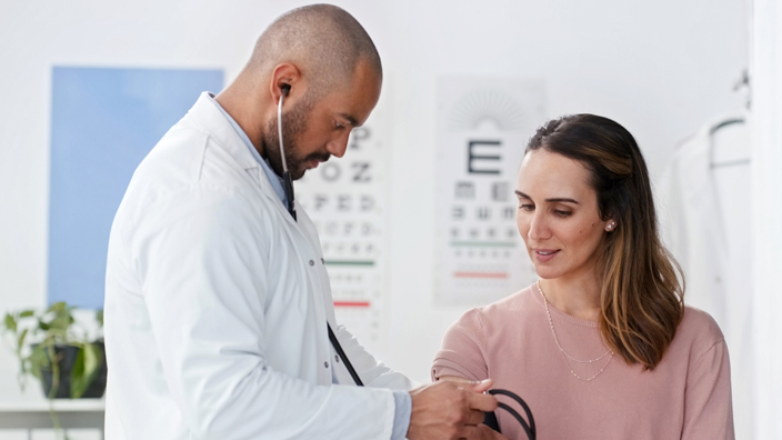 A male doctor wearing a white coat speaks with with a female patient wearing a pink shirt