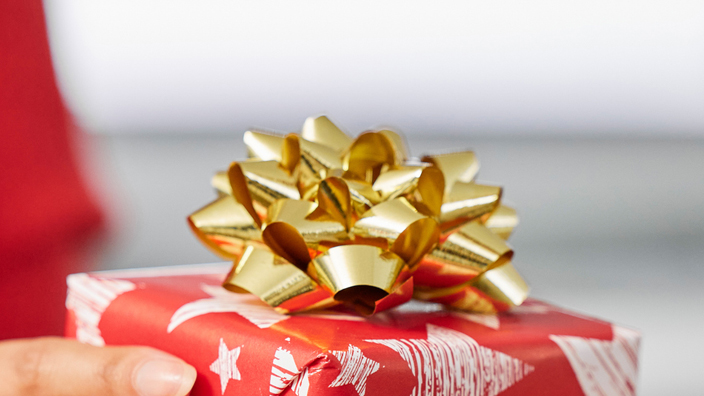 One person hands a gift wrapped in red paper and adorned with a gold bow to another person