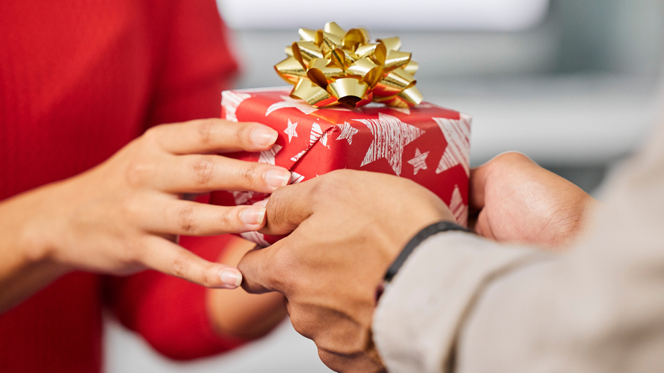 One person hands a gift wrapped in red paper and adorned with a gold bow to another person