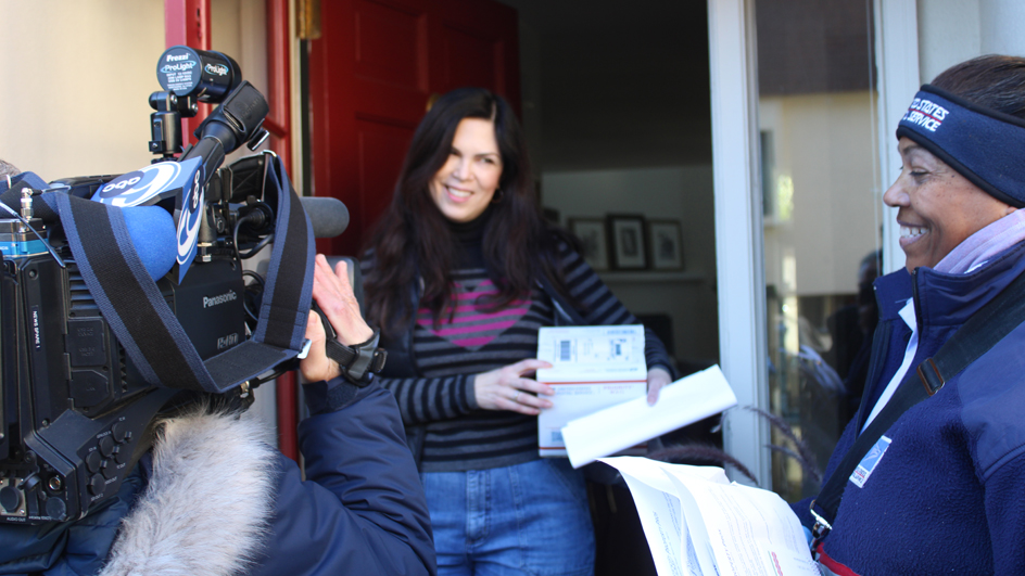 A woman receives a package at her front door from a USPS letter carrier.