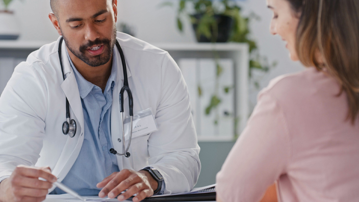 A male physician wearing a white lab coat and stethoscope talks with a female patient.