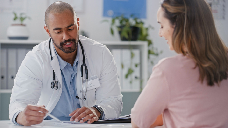 A male physician wearing a white lab coat and stethoscope talks with a female patient.