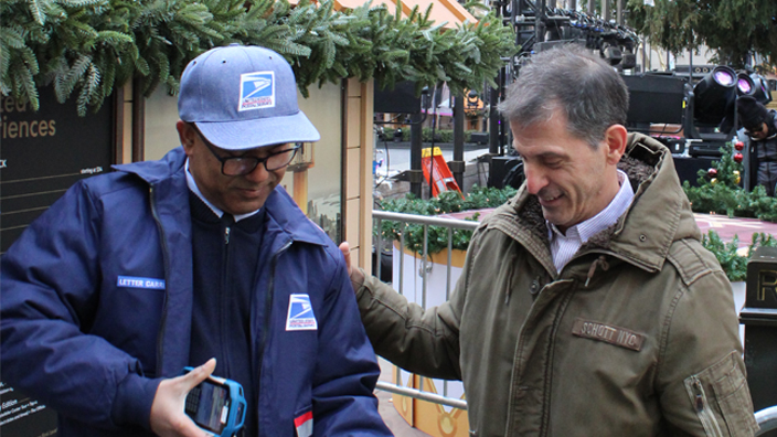 A USPS letter carrier scans a package with a handheld device before handing it to a customer.